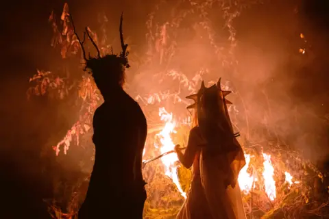 Kitt Bunney A man and a women are stood in front of a fire and we can see their silhouette. The man has horns coming from his head and the women has a form of veil on.