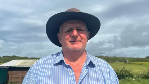 A man wearing a brown sunhat and a blue and white striped shirt stands in front of a field with a shed visible behind him 