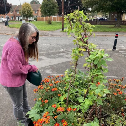 A woman pours water from a watering can into a flowerbed which is full of plants and flowers. 