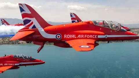 A close-up aerial image of Red Arrows aircraft, flying over the blue sea in close formation. One has red, white and blue smoke emerging from the back.