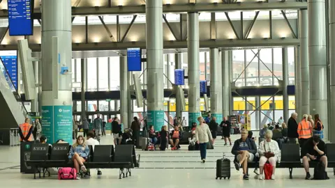 Pacemaker The foyer area of the new station. Passengers are sitting in seating areas waiting for their buses. Some have suitcases and travel bags.