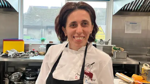 Denisa Theodorescu stands in front of a window in a kitchen in chef's whites with a black apron over the top. She has thick, wavy dark hair and is smiling