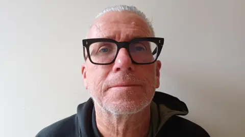 Richard Osborne A man with short, neatly-combed grey hair and a well-groomed white beard. He is wearing large, black-framed glasses. His outfit consists of a hooded sweatshirt in olive green with contrasting black sleeves. The background is a plain, light-coloured wall.