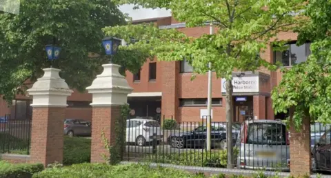 Se puede ver la entrada a la comisaría de Harborne, con un portón de ladrillo a la izquierda y farolas azules de la Policía de West Midlands sobre dos pilares. Más allá se ven árboles en un aparcamiento con varios vehículos estacionados frente a la entrada principal de la comisaría.