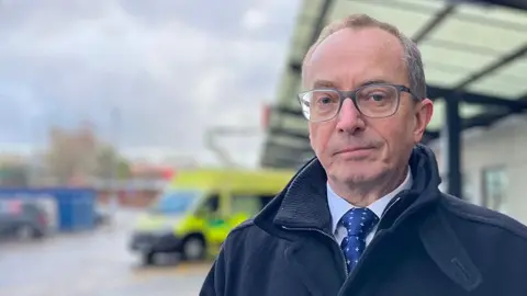 Dr Jim Gardner, chief medical director of Liverpool University Hospitals NHS Foundation Trust. He has glasses and is photographed outside Royal Liverpool University Hospital while wearing a dark blue tie, pale blue shirt and a navy blue coat. Out of focus, an ambulance is behind him.