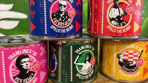 Simon Thake/BBC Five miniature multi-coloured cans are stacked together in a pyramid shape, showing pictures of musicians set to play at an upcoming festival.