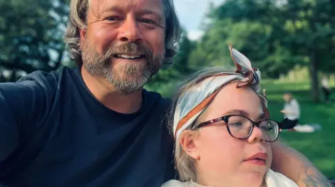 Darren Scott Darren and Sophia Scott. He has longish blond hair and a beard and is wearing a blue T‑shirt. His arm is around Sophia who is wearing glasses and a blue, brown and white cotton scarf tied around her head. He is smiling at the camera while she is looking off camera. They are in a park.