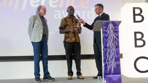 Mark Pemberton A man receives an award on stage, in front of a screen. There is a large light-up sign that reads BBC. There is a purple podium. A man is holding a microphone to the award recipient, and another man stands and watches