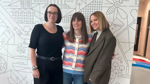 Hannah Harrison, Annette Illing and Kimberley Walsh are standing side by side and are smiling at the camera in this posed photograph. 