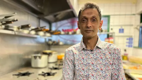 Syad Ahmed wearing a floral shirt. He is inside a restaurant kitchen with pots behind him. He has brown hair and eyes.