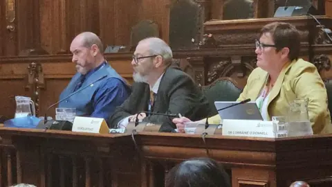 Aisha Iqbal/BBC A panel of three people sitting at the head of a meeting in a council chamber, two men and one woman.