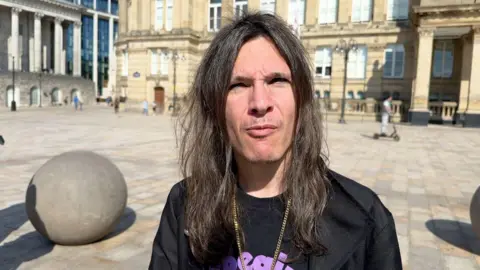 A close-up of a man with long dark hair, wearing a Black Sabbath shirt and a gold chain.