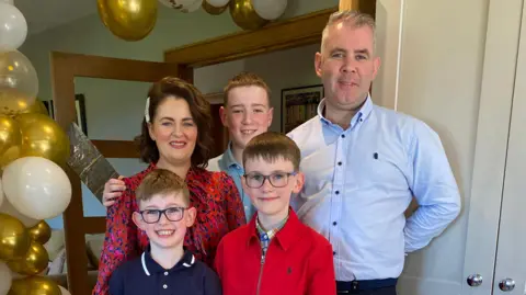 Fergal Sherry The Sherry Family all stand and pose for a picture in a room with gold and white balloons at the side. Catherine is stood on the left and is wearing a red, blue and pink leopard print dress and has short, shoulder-length brown hair. Beside her is her eldest son who has a blue shirt on and has short light coloured hair. On the right is Fergal, who has short grey hair and is wearing a shirt collared shirt. At the front are the two youngest sons, who both have medium-length brown hair and are wearing dark framed glasses. All of them are smiling at the camera.