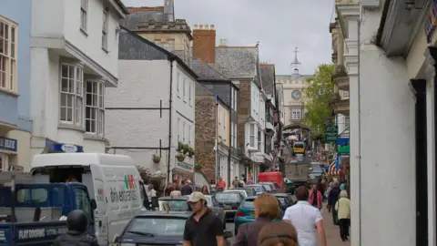 A busy shopping street in Totnes with cars and vans heading up the road under a cream arched gatehouse which has a clock face on it. There are lots of people walking up and down Fore Street.