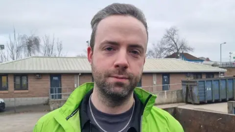 Man with grey hair wearing a bright green jacket, grey shirt and silver necklace, pictured in front of a red brick single storey building