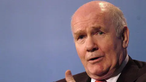 Close up shot of John Reid talking as he gestures with his hands.