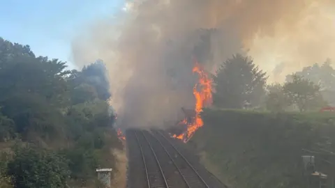 Flames tower above a large hedgerow on the right side of a railway line. A huge plume of smoke completely blocks the view up the line.