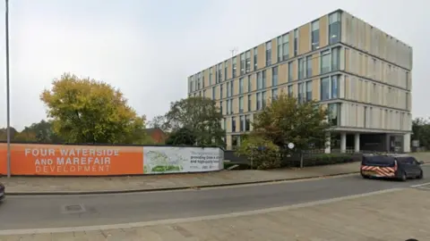 Google An orange and white banner, with the words "Four Waterside and Marefair", is seen alongside a town centre road. There are trees behind the banner.  To the right is a light brown and grey modern building with five storeys - the University of Northampton Innovation Centre.