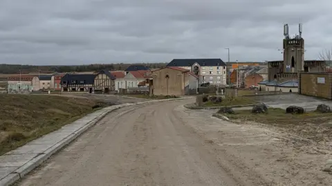 A wide view of the simulated village with two to four-storey buildings and a church-type tower looking above on the right. A road leads down to the village.