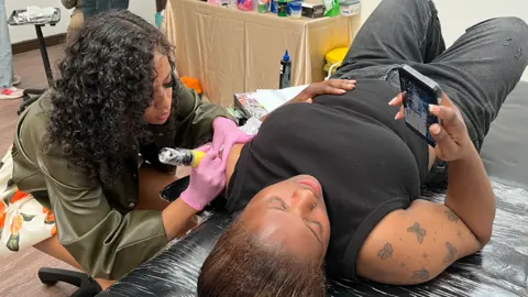 A woman in a black top lying on a tattooist's table as the tattoo artist on the left creates an image on the top of woman's left shoulder. There is a workstation in the background, covered in a gold-coloured cloth, with various products on it