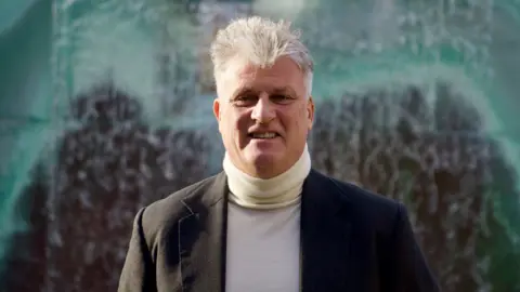 A man with grey hair stood in front of a water cascade in front of a green and brown wall. the man is wearing a white polo neck jumper and grey jacket.
