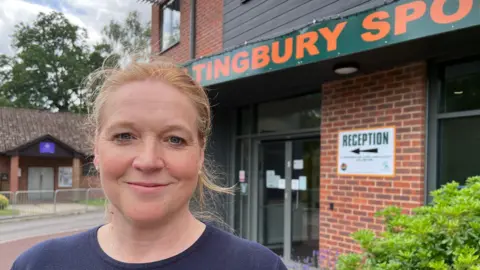 Lady with tied black red hair and a blue jumper stands in front of a two storey brick building. The sign says reception and you can read part of the Hiltingbury sports sign.