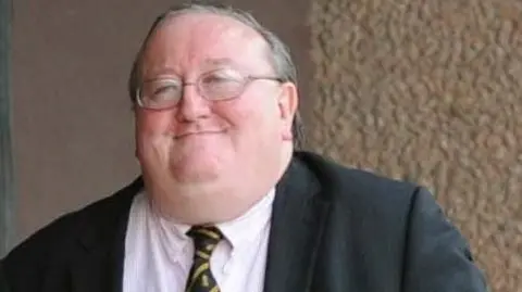 George Gordon is pictured with short grey hair and glasses. He is wearing a black blazer, a light pink shirt and a black and yellow tie