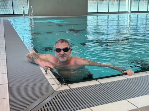 A man with dark hair and dark goggles is in the pool at one edge, with his arms up over the side