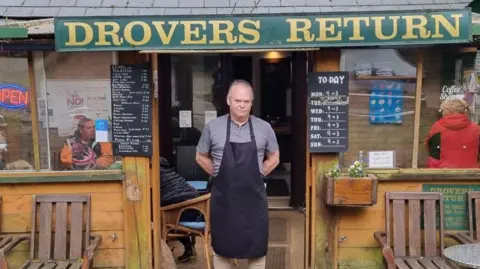 A man, standing in the doorway of a cafe, wearing a black apron, and a grey top. People are inside the building. There are signs above and either side of the door and chairs and a table outside. The man looks serious. 