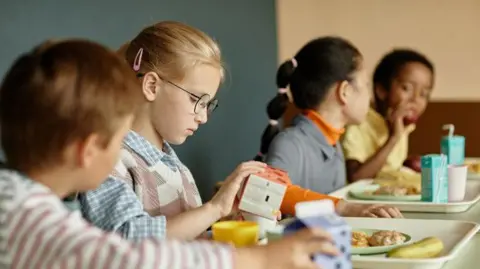 Getty A blonde-haired child with black round glasses pours orange juice into cup. She is sitting on school dinner table with three other children who are also eating. 