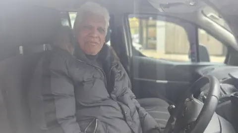 Aisha Iqbal/BBC A grey-haired man of South Asian sits in the driver's seat of a car. He wears a puffy black jacket with a fur lined hood.