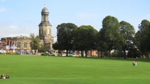 Shrewsbury Town Council A landscape photo showing a mother and child walking across a large patch of grassland. Two people are sat down to the left of the photograph. In the distance a line of trees leads up to a built up area with an historic looking building with white and black beams on the front of it. A very large building behind it has arched windows and a tall tower with a dome shape on the top. At the top is a crucifix. 