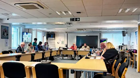 Dr Simon Desert addresses members of Cumberland Council's health overview and scrutiny committee. In total, about eight people are sitting at wooden desks arranged in a rectangle shape. Each member has a microphone in front of them.