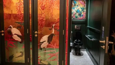 A black toilet on the right in a toilet cubical. On the left there is a red and yellow tropical design with flamingos on the cubical doors.