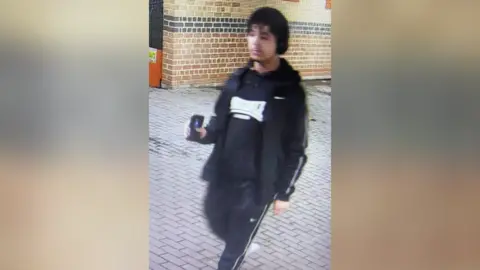 CCTV image of a young man in a black Nike tracksuit and black hoody, holding a phone in his hand.