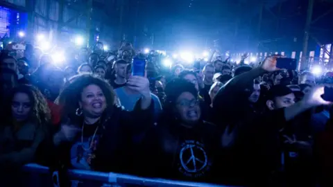 A large group of people holding up their phones inside Drumsheds nightclub.