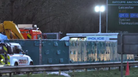 A fence with dark green screens is in place at the scene, as well as two recovery vehicles and a police van