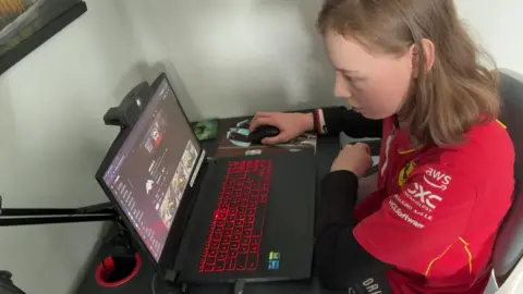A teenager with long blonde hair sits at a laptop with red lit keyboard keys, looking at YouTube videos. He's wearing a red Ferrari t-shirt with long black sleeves underneath
