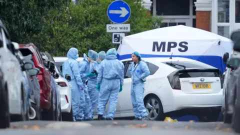 PA Media Forensic officers in blue protective suits gather at a crime scene on a residential street next to a white Toyota Prius and a blue and white Metropolitan Police Service (MPS) tent.