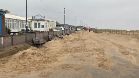 BBC News The photo shows a beach promenade. It is covered with piles of sand on either side. There are five benches that are also covered. Along the left hand side, behind a wall, are a row of shops. On the right hand side are some railings which lead onto the sea.