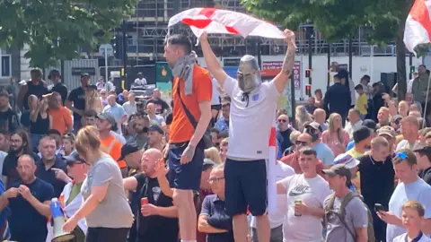 PA Media Two men stand on a raise platform in the centre of a far-right demonstration. One is wearing an England football shirt, with an England flag and a silver knight mask, while another is wearing an orange shirt and blue shorts. A group of people are behind them, with one woman carrying a megaphone