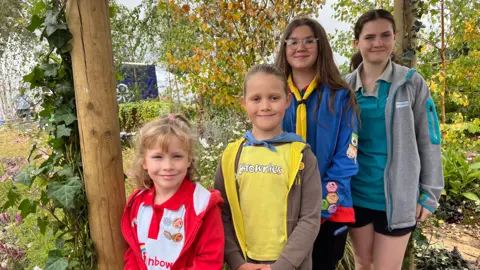 Four girls stand in a garden with lots of trees with green and yellow leaves behind them. The smallest child is wearing a red outfit. Next to her is a slightly taller girl wearing yellow. Next to her is a taller girl wearing dark blue and on the end is another slightly taller girl wearing a blue t-shirt and grey hooded top. 
