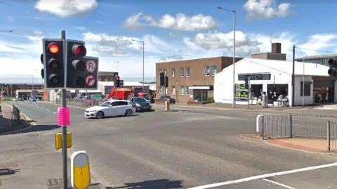 A white car crosses a road junction as a dark vehicle waits by traffic lights on red