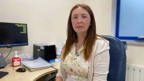 Dr Michelle Le Cheminant is wearing a white dress with patterned pale flowers and a creme cardigan. She has medium length light brown straight hair.