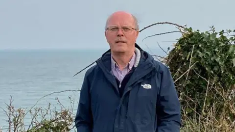 Maurice Parkinson A grey haired man wearing square-shaped glasses and a navy North Face coat looks at the camera while standing in front of some shrubbery and water