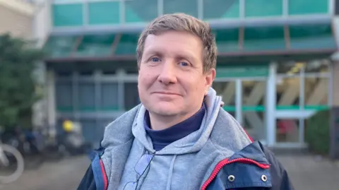 A fair-haired man wearing a grey hoodie and blue jacket standing in front of a building with a glass frontage. Parked bikes can be seen over his left shoulder.