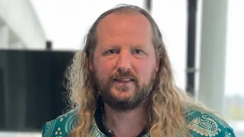 Edd Smith/BBC Finbarr Carter is in the middle of the frame. He has long blonde hair and is wearing a blue and cream patterned shirt. He is standing in front of a large glass window. 