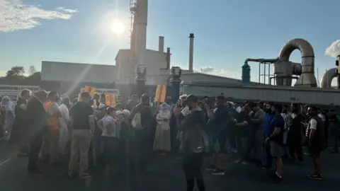 Aisha Iqbal/BBC A large group of people, many wearing masks, standing outside a factory. It's a mixed crowd of men, women and children. Large metallic pipes and industrial structures behind the crowd identify the location as a factory or processing plant of some sort.