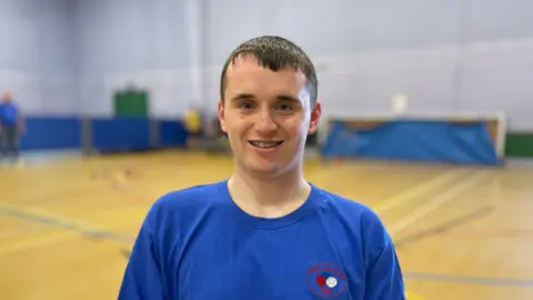 Luke sits in the middle of a gymnasium. He is wearing a blue top with the red and white GEM Boccia logo on it. He is smiling at the camera. 