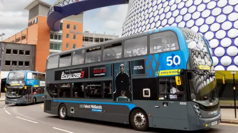 A National Express bus drives past part of the Bullring Shopping Centre in Birmingham with a second bus behind it.